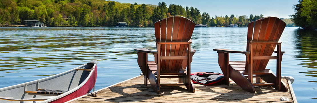 Two Muskoka chairs on a wooden dock overlooking the blue water of a lake in Muskoka, Ontario Canada. A red canoe is tied to the pier and life jackets are visible near the chairs.